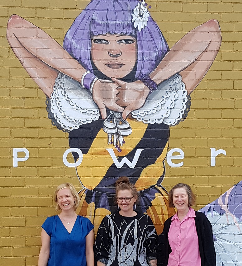 Photo of three women below a mural of a woman with word POWER written across her chest