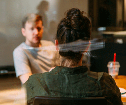 Two seated people talking, a woman with her back to the viewer and a man